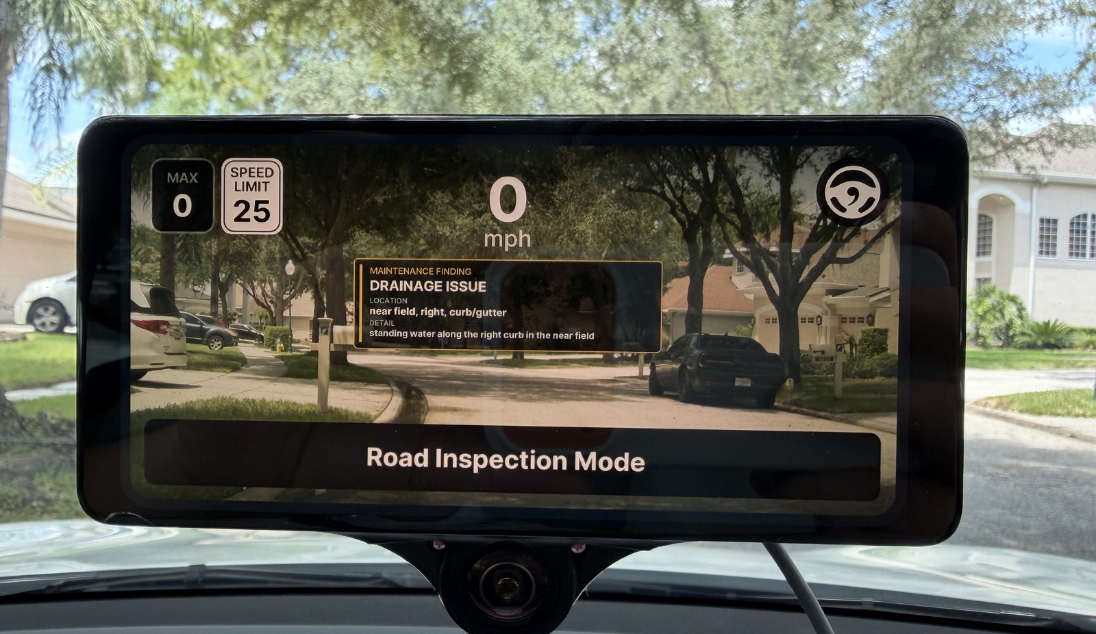 A windshield-mounted DoTPilot unit running in Road Inspection Mode. The display shows the forward camera view of a residential street overlaid with a maintenance finding card reading "DRAINAGE ISSUE — location: near field, right, curb/gutter — detail: standing water along the right curb in the near field," alongside the current speed and posted speed limit.